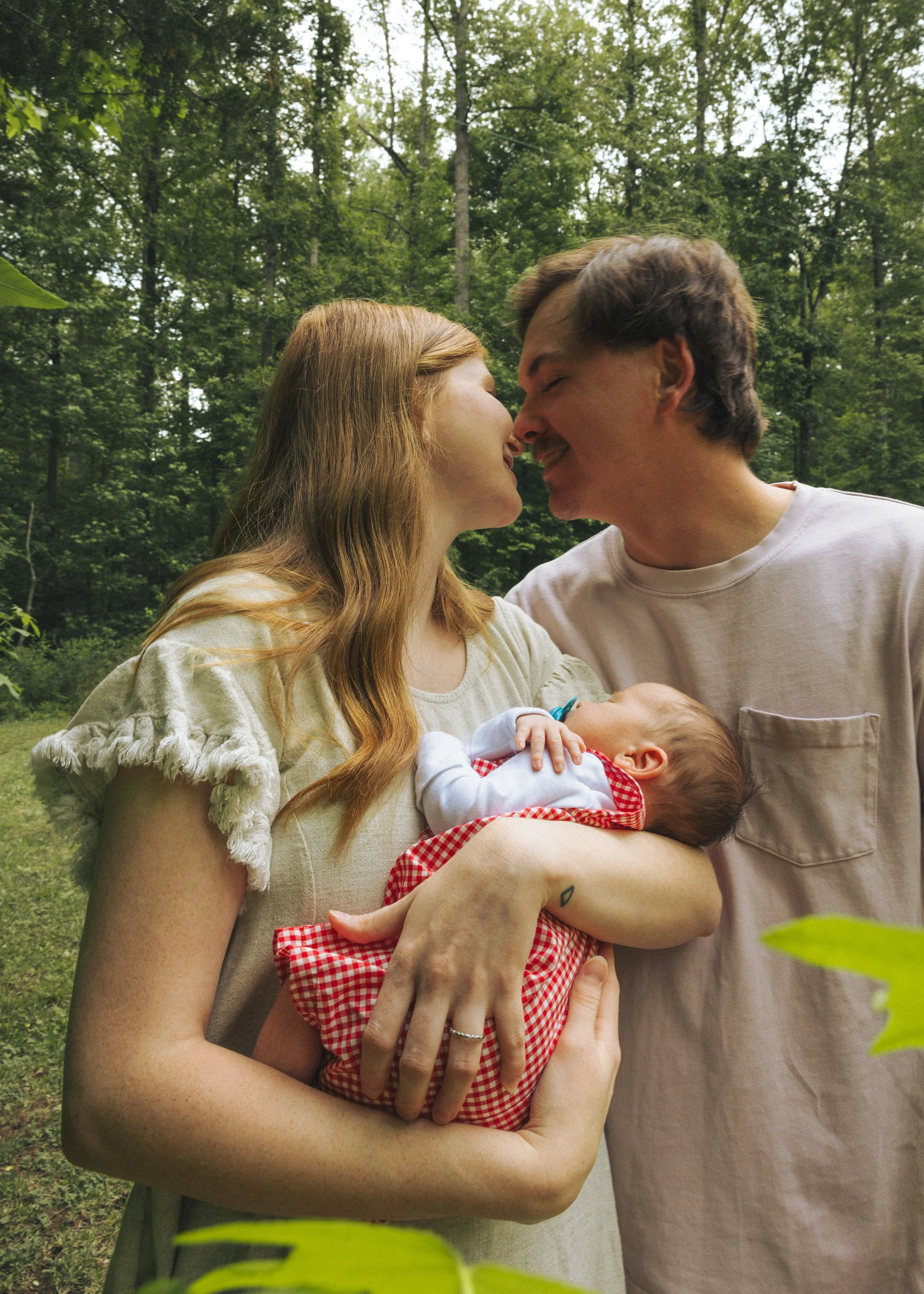 A woman holding a baby outdoors with a man leaning in and touching noses in front of a wooded background.
