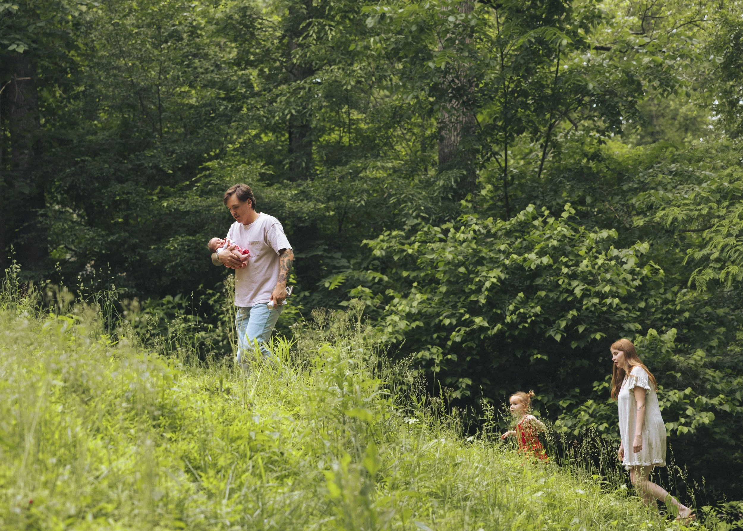 A woman holding a baby in a forest clearing with two young girls nearby, surrounded by green trees and foliage.