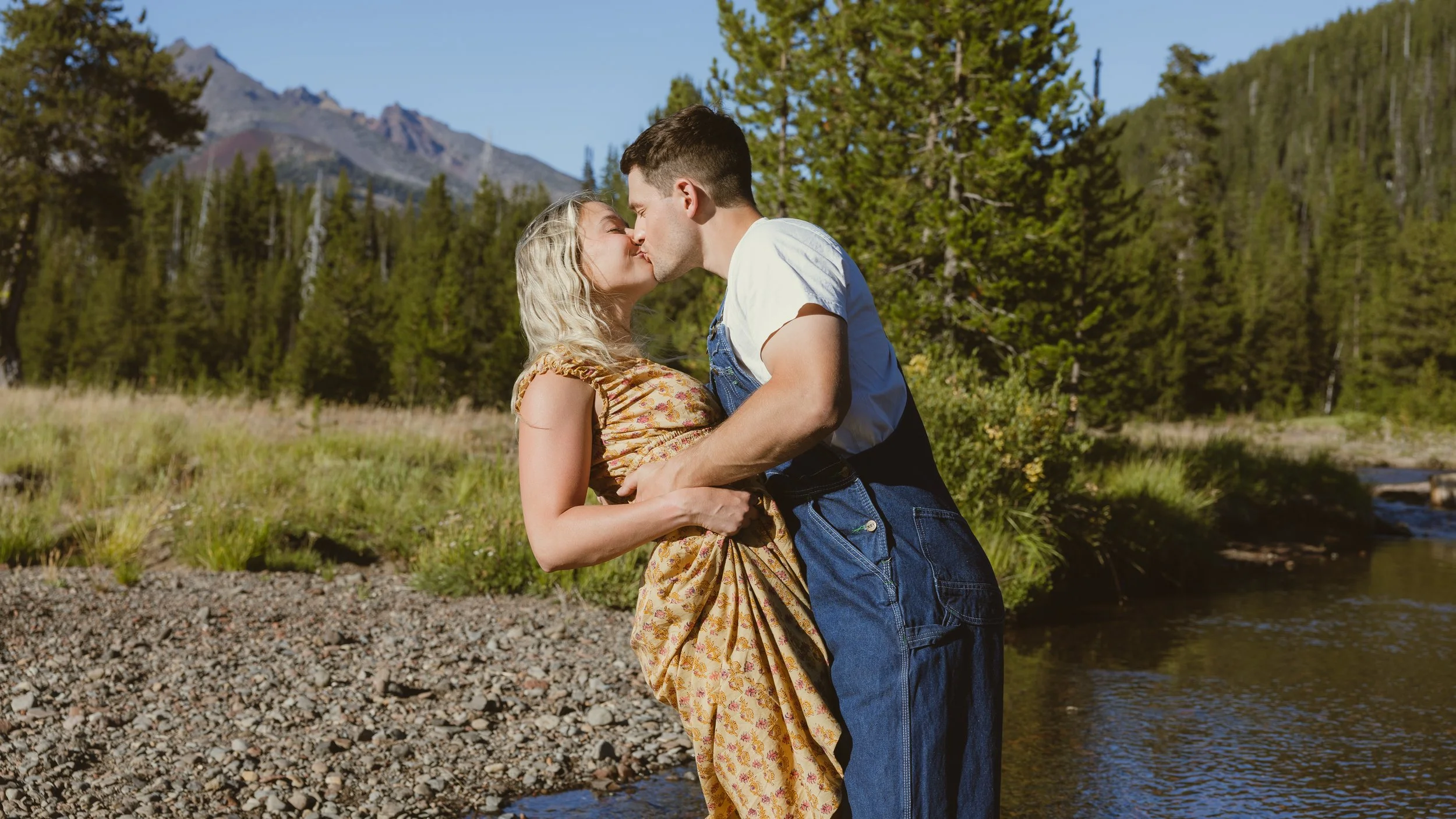 A couple kissing outdoors near a river, surrounded by trees and mountains in the background.