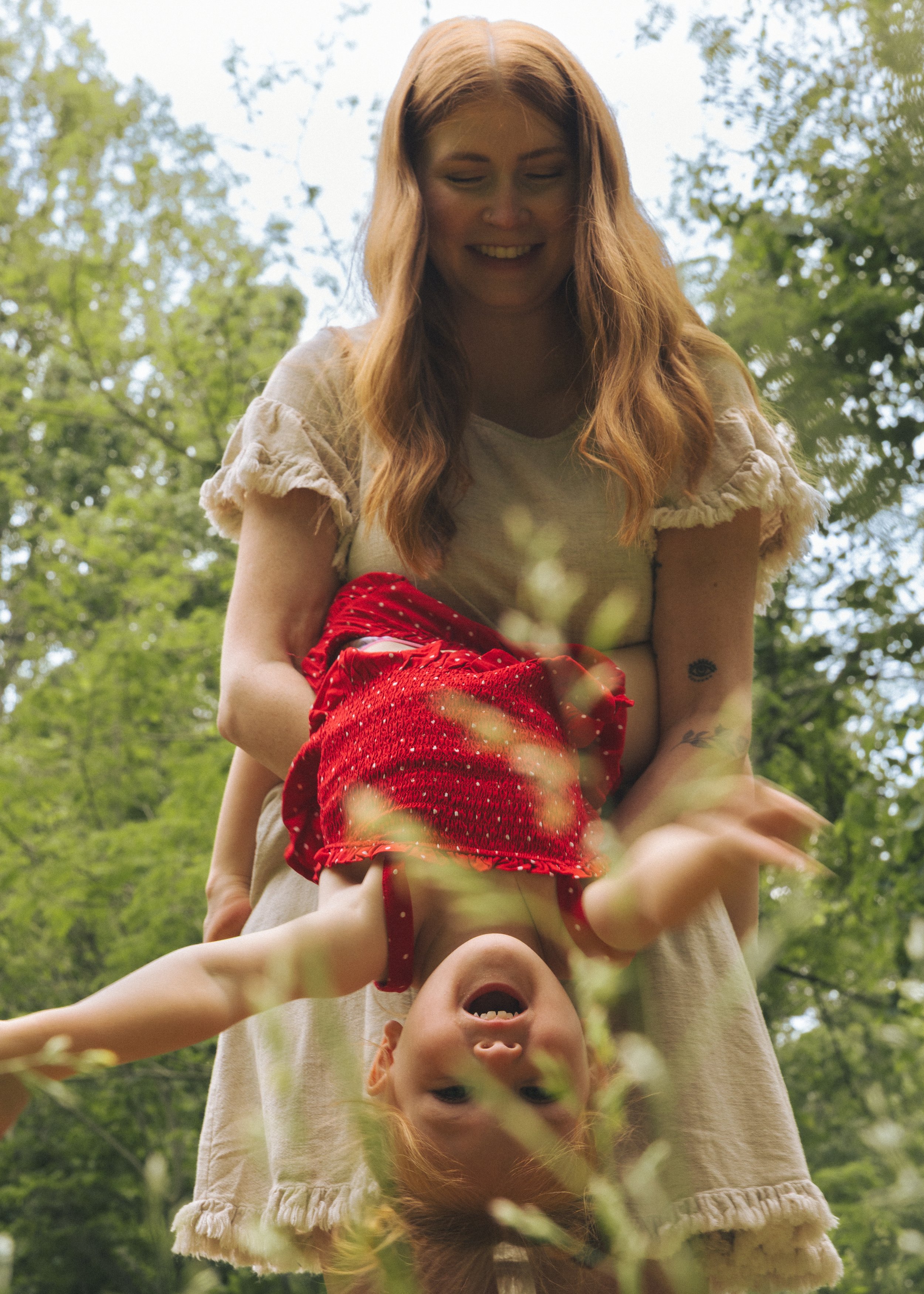 A woman with long red hair and tattoos on her arm is giving a piggyback ride to a young girl with red hair in a green outdoor setting with trees.