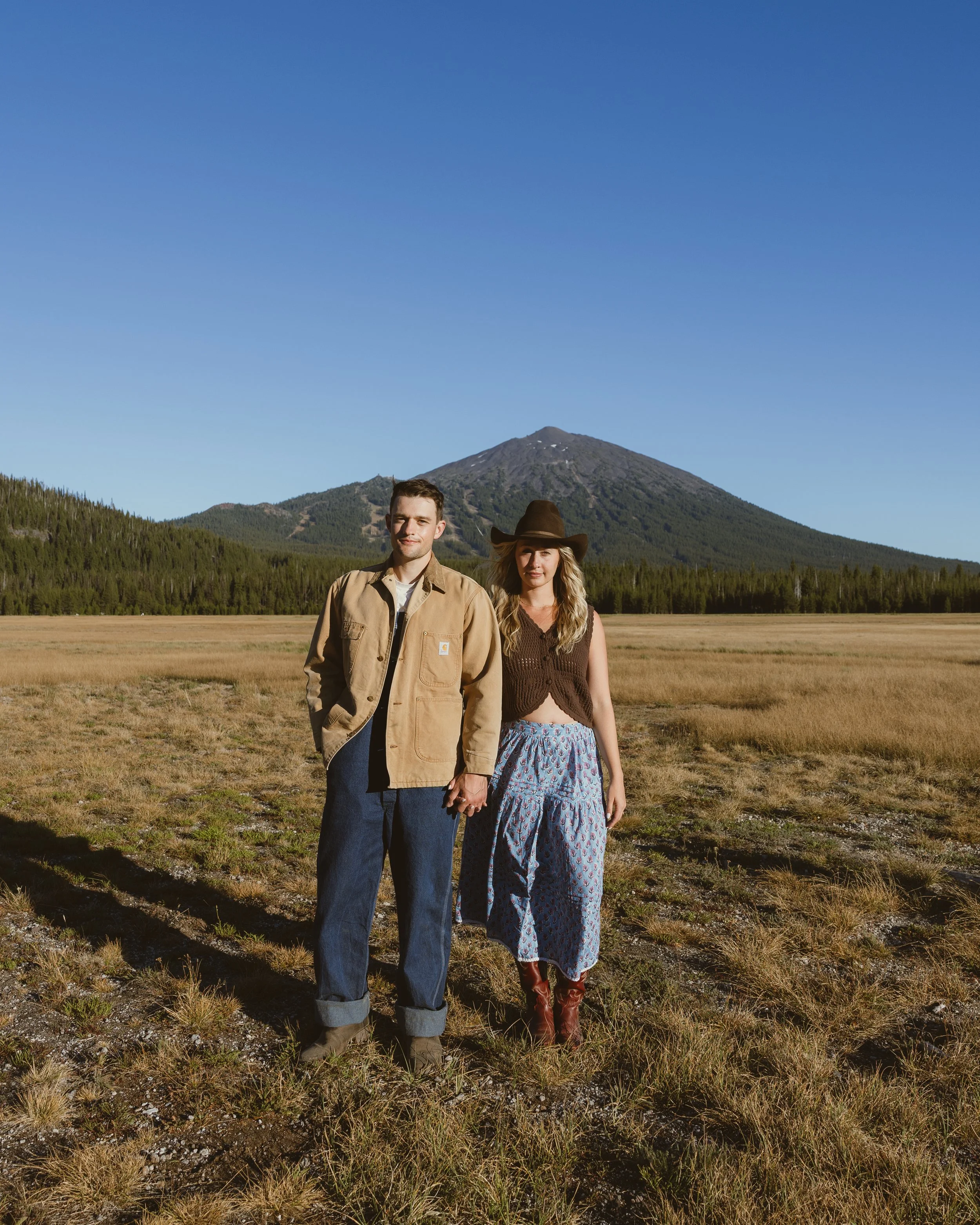 A young man and woman holding hands in a field with mountain in the background. The man wears a tan jacket and blue jeans, and the woman wears a brown top, patterned skirt, cowboy boots, and a wide-brimmed hat.