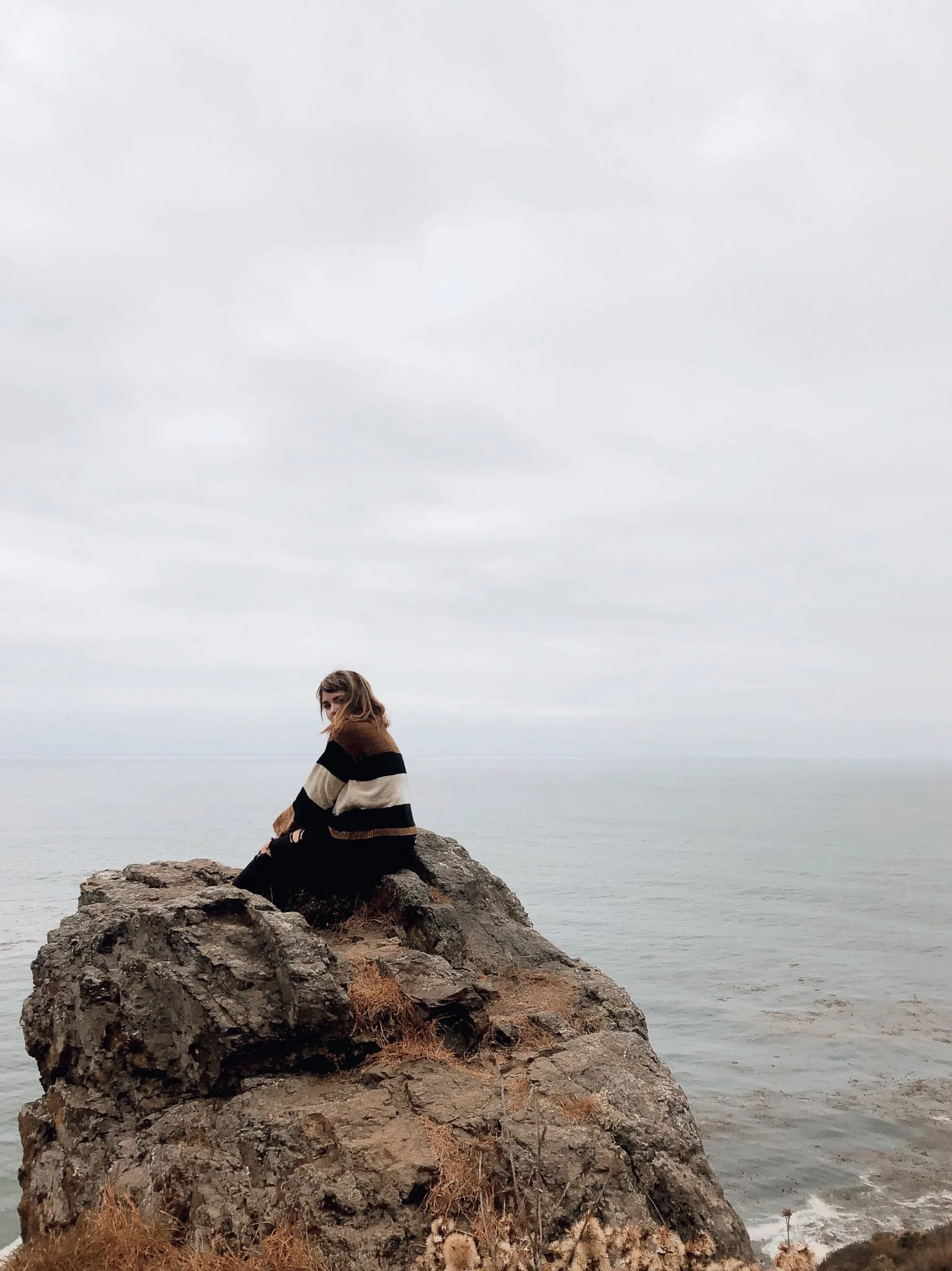 A woman sitting on a large rock by the ocean on a cloudy day.