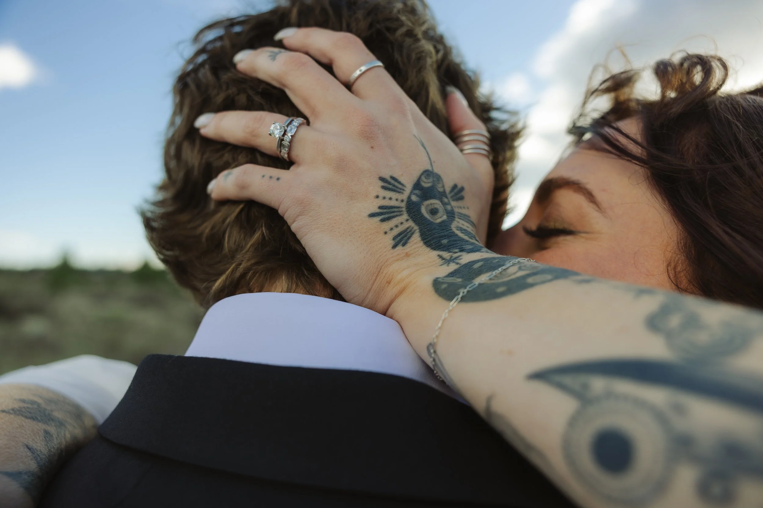 Close-up of two people embracing outdoors, with one person's hand on the other's head showing rings and tattoos. The background is a cloudy sky.