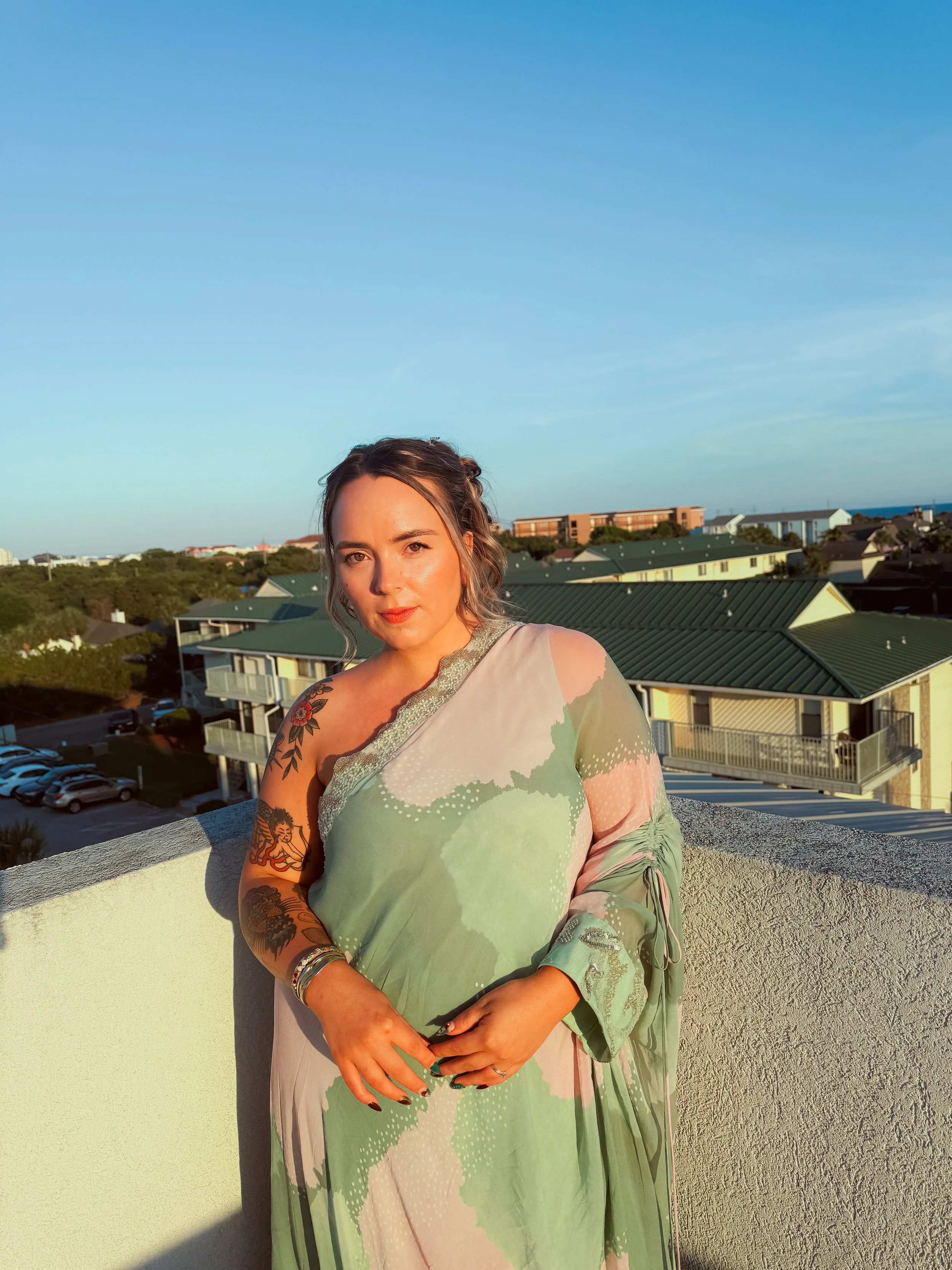 A woman with tattoos on her right arm and dark hair styled in loose waves stands on a balcony during sunset, with residential buildings and the ocean visible in the background.