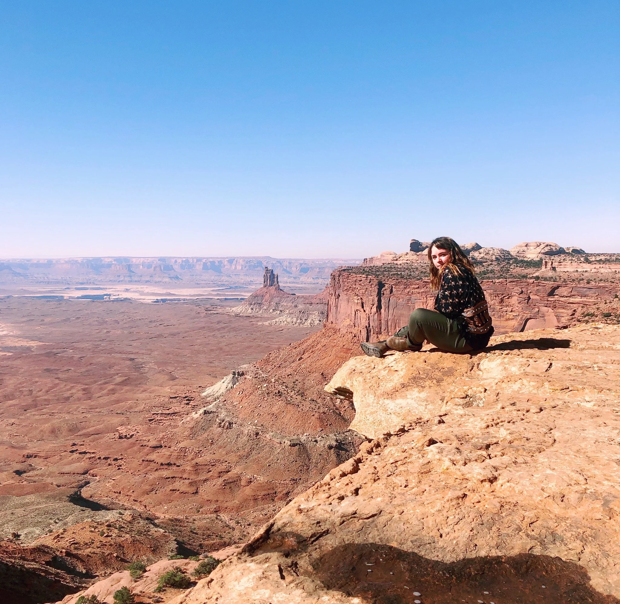 A young woman with brown hair and a black floral jacket sitting on a rocky ledge at the Grand Canyon during daytime. The expansive canyon landscape stretches into the distance with layered rock formations and a clear blue sky overhead.