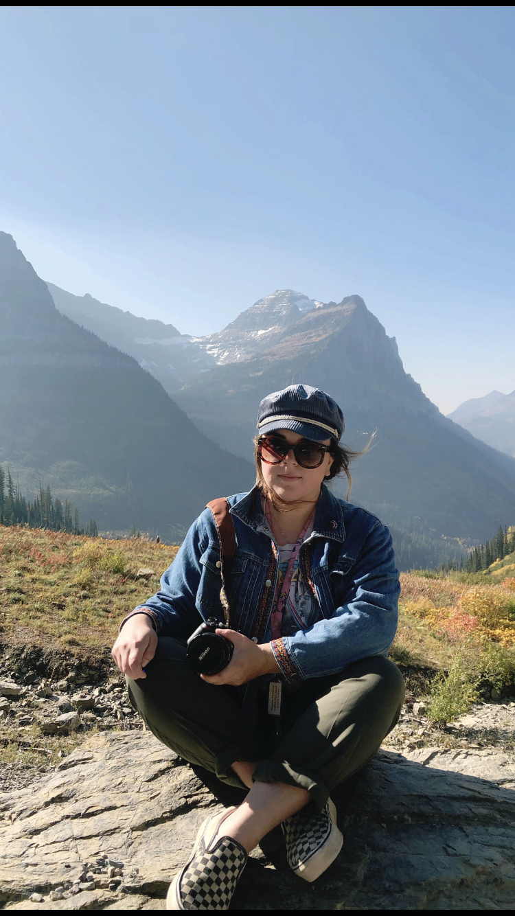 A woman sitting on a rock outdoors in a mountainous landscape, holding a camera, wearing sunglasses, a cap, a denim jacket, and checkered shoes.
