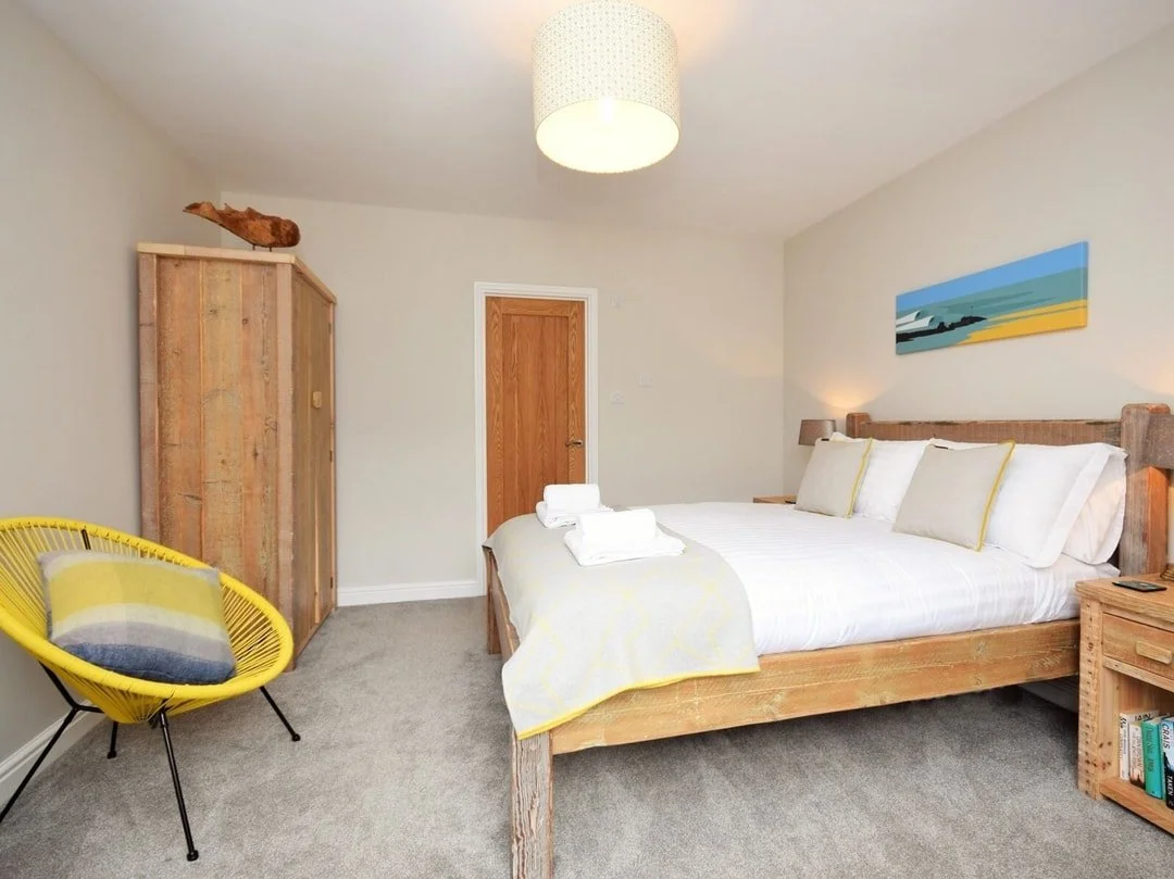 A bedroom with a wooden bed, white bedding, and pillows, a wooden wardrobe with a decorative object on top, a yellow chair with a pillow, a nightstand with books, a wall painting, and a ceiling light.