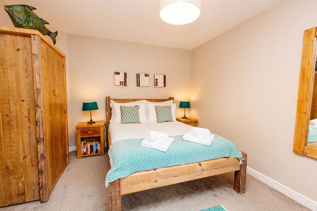 A cozy bedroom featuring a wooden bed with white bedding, two green pillows, and a teal blanket. Two wooden nightstands with teal lamps flank the bed. An armoire and a large mirror, both made of wood, are also in the room. There are three small frame