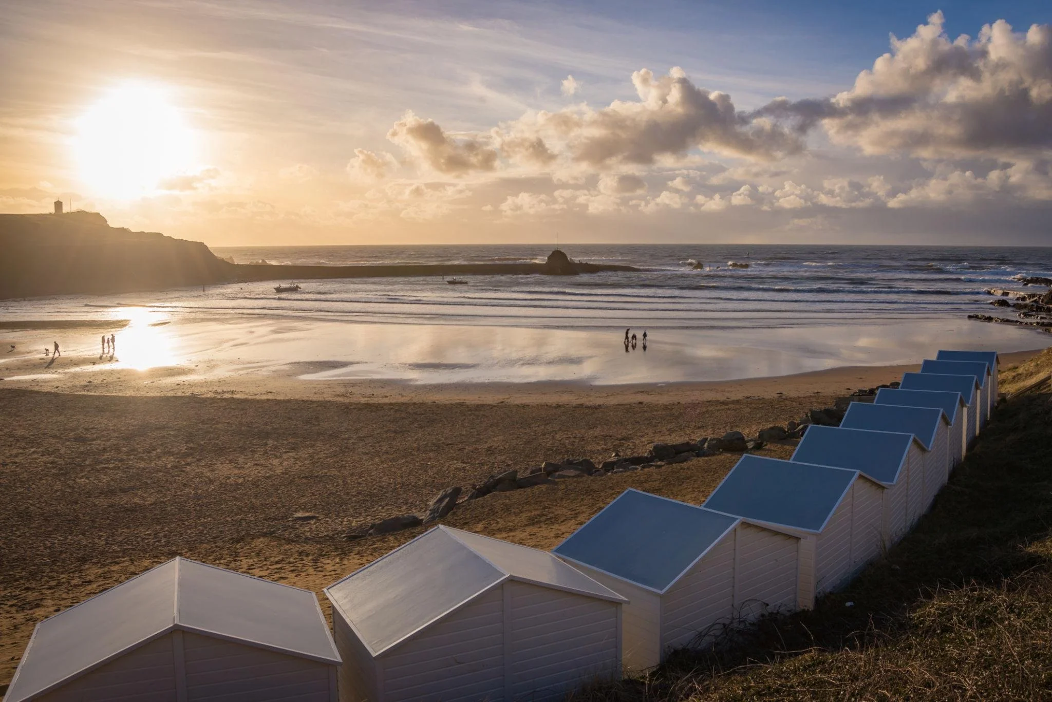 Sunset over a beach with small white beach huts in the foreground and people walking along the shoreline.