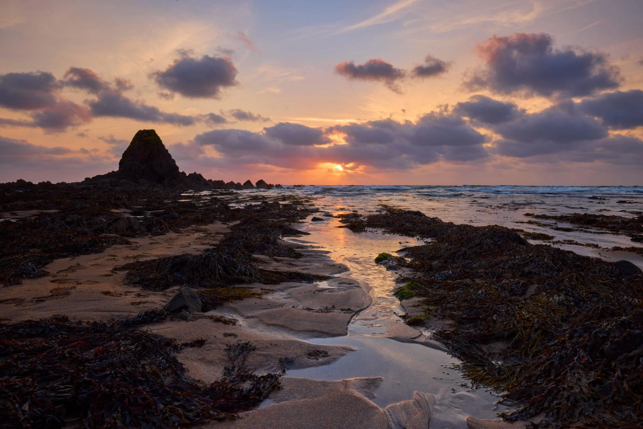 Sunset over a rocky beach with a large rock formation in the distance, colorful sky with clouds, and tide pools reflecting the sunset.