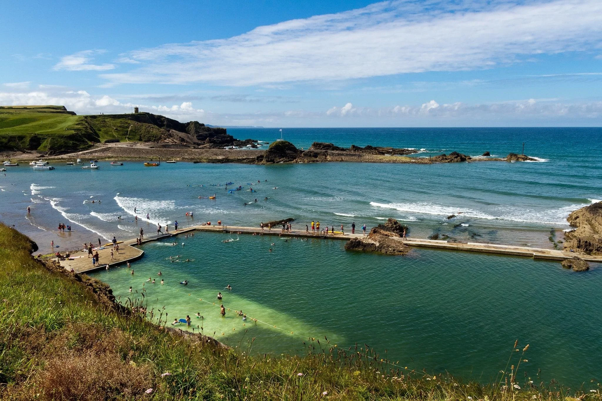View of a beach with people swimming and lounging in the water, boats anchored nearby, green hills in the background, and a partly cloudy sky.