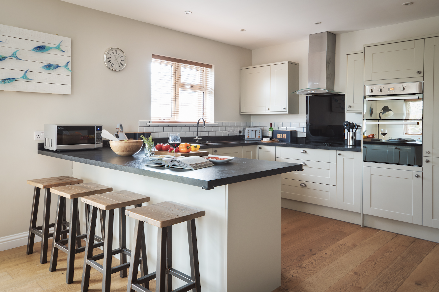A modern kitchen with cream-colored cabinets, black countertops, and a white subway tile backsplash. There is a kitchen island with wooden stools, a microwave, a bowl of vegetables, a glass of wine, and an open cookbook. The kitchen has a window with
