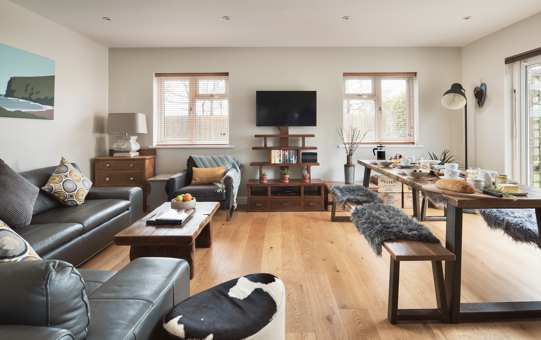 Living room with gray leather couch, wooden coffee table, and armchair with colorful pillows. Flat-screen TV mounted on wall, wooden entertainment unit, and window with blinds. Dining table set for breakfast with bread, bowls, cups, and jars. Wood fl