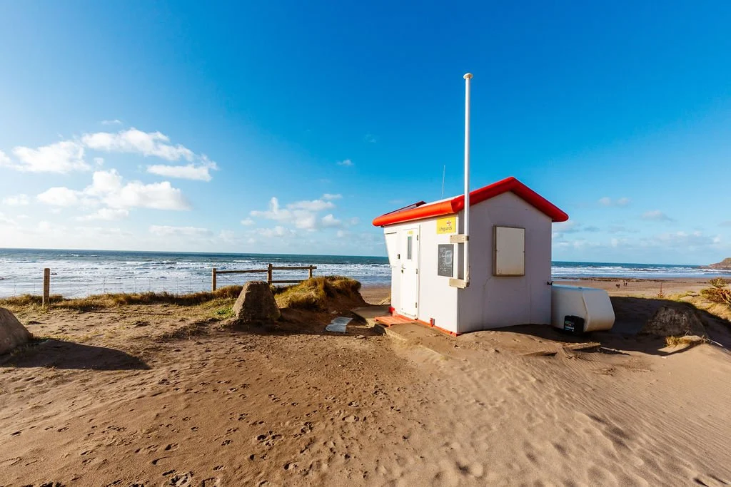 Small beach lifeguard house with a red roof on sandy dunes, overlooking the ocean under a bright blue sky with a few clouds.