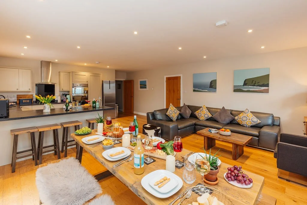An open-concept living space with a dining area and kitchen. The dining table is set with plates, glasses, and various food and drinks. The living area has a dark sectional sofa with decorative pillows, a wooden coffee table, and artwork on the walls