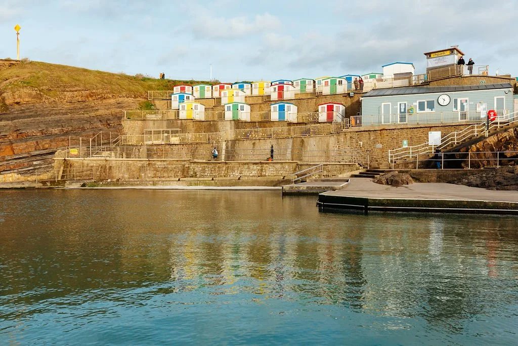 Colorful beach huts on a hillside overlooking a body of water, with a small building and a clock on a gray structure at the shoreline.