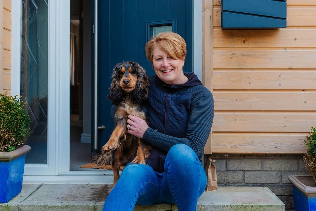A smiling woman with short red hair sitting on the doorstep of a house, holding a brown and black dog.