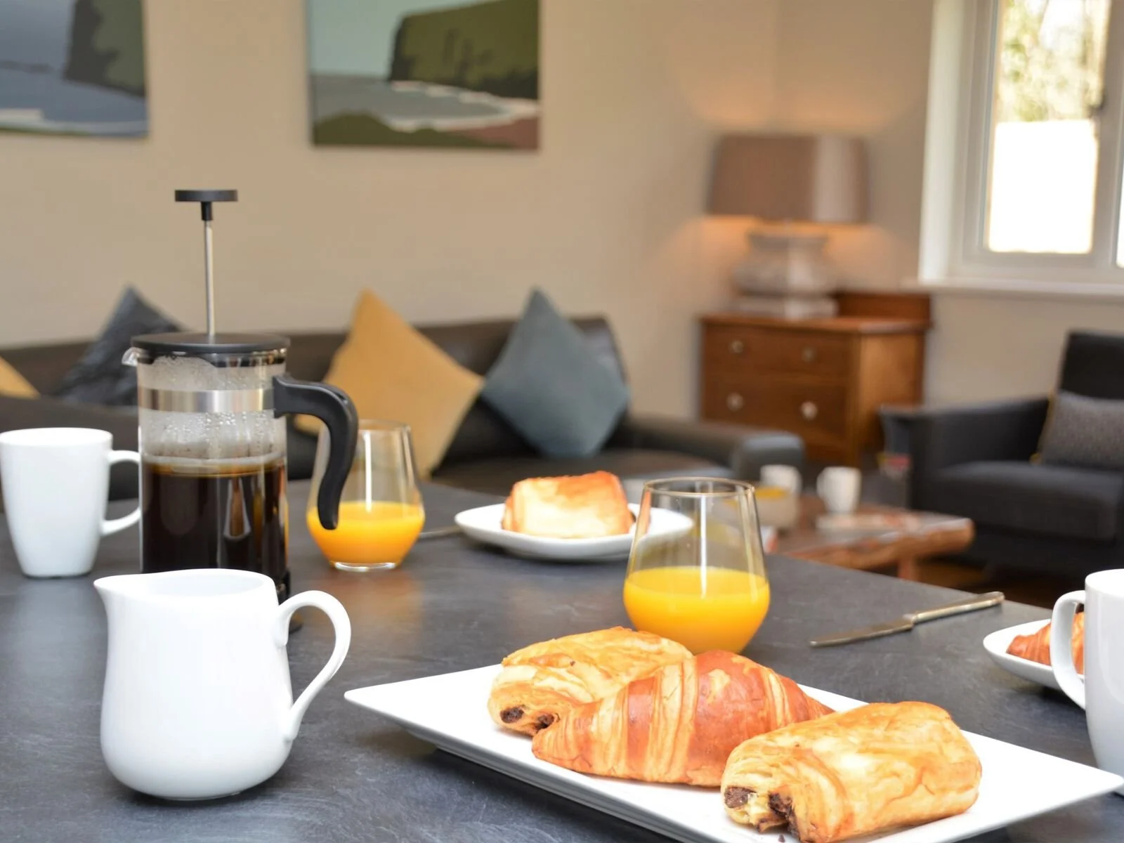 A breakfast scene on a dining table with croissants, orange juice, coffee, and a French press, set in a living room with a sofa, pillows, and a wooden side table.