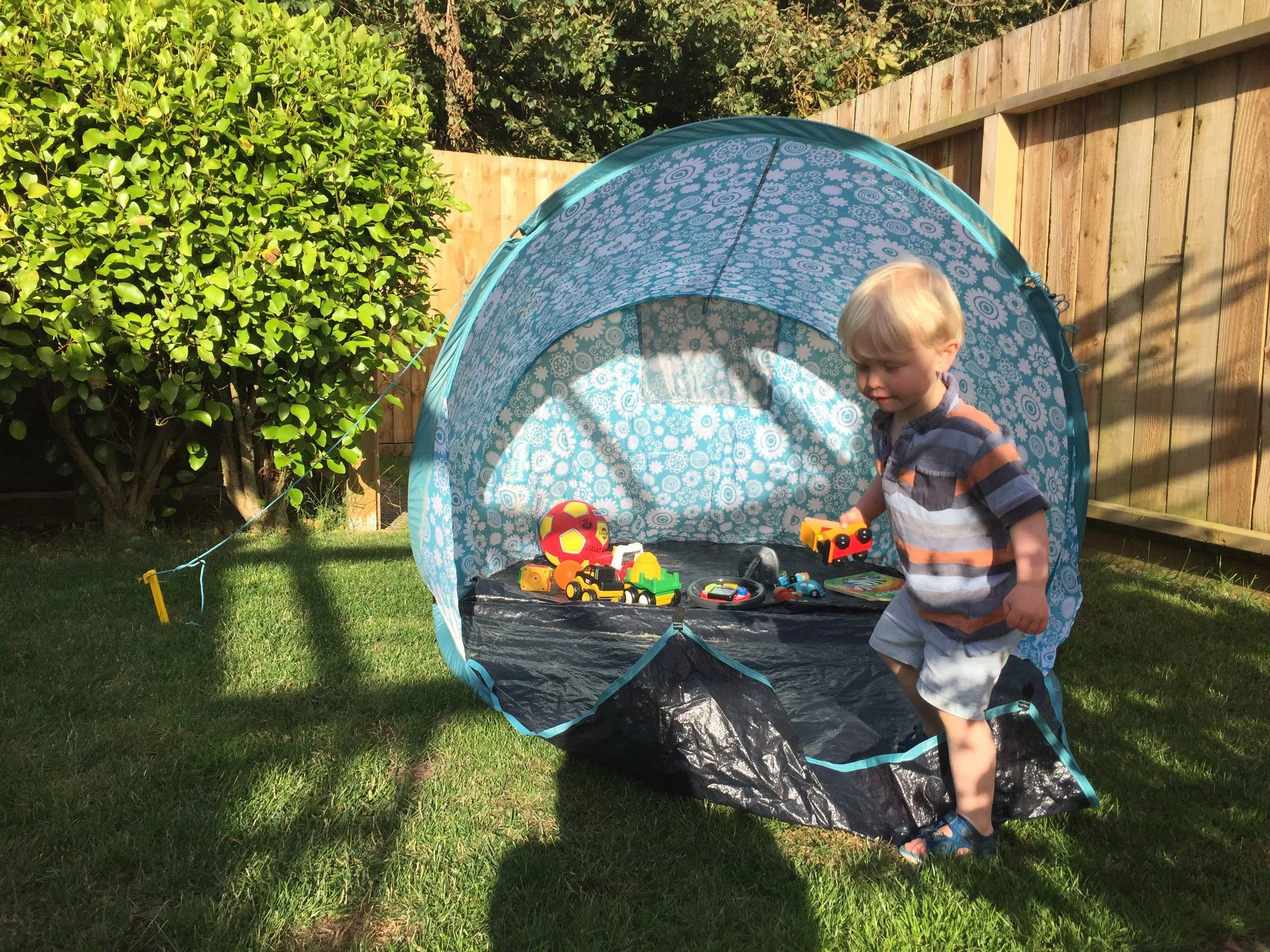 A young boy with blonde hair wearing a striped shirt and shorts playing outside in a yard with a small blue pop-up tent filled with toy cars, a ball, and other toys, with a wooden fence, green bush, and trees in the background.