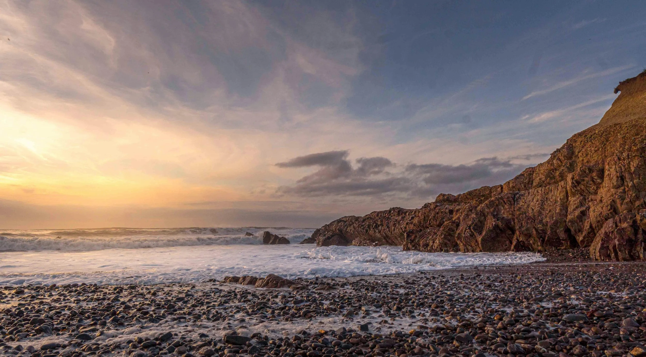 A rocky beach scene at sunset with waves crashing, a large cliff on the right, and a partly cloudy sky.