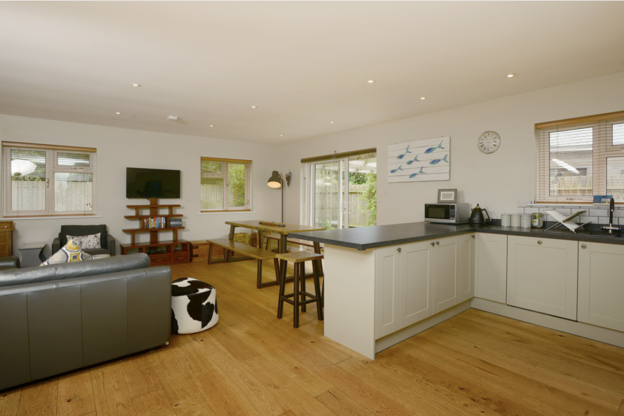 Open-concept living room and kitchen with white walls, hardwood floors, and modern furniture including a gray sofa, black and white cow-print ottoman, and wooden dining table with benches. The kitchen has white cabinets, a black countertop, a microwave, and a window above the sink. Recessed ceiling lights and a sliding door lead to the backyard.