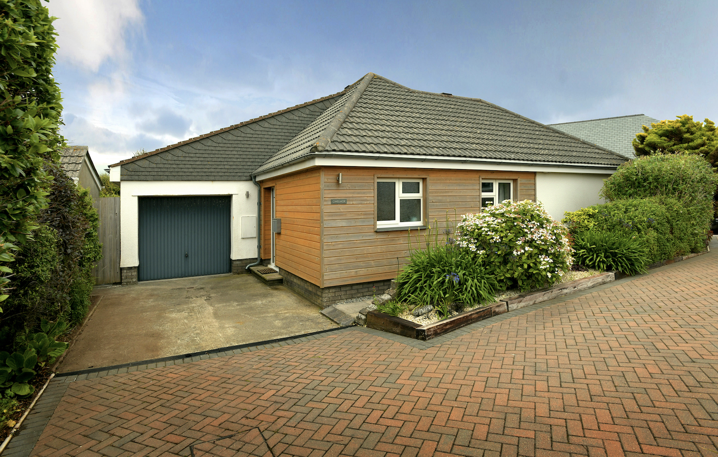 Residential house with a garage, surrounded by landscaped plants and a brick paved driveway under a partly cloudy sky.