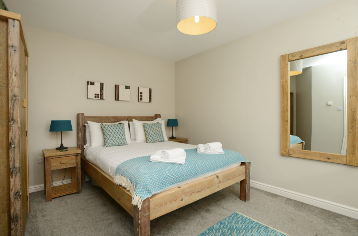 A bedroom with a wooden bed, white bedding, and blue decorative pillows. There are matching blue lamps on wooden nightstands on either side of the bed. A large mirror with a wooden frame hangs on the wall to the right. The room has light-colored walls and carpet.