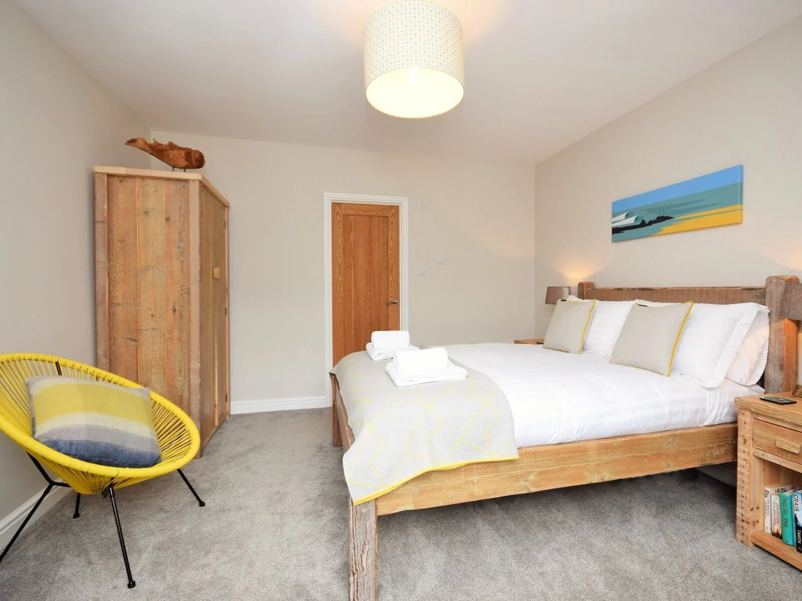 A bedroom with a large bed, white bedding, four pillows, and two rolled towels. There is a wooden wardrobe on the left, a yellow accent chair with a pillow, a wooden door in the background, and a colorful landscape painting above the bed. A bedside table with a lamp is on the right.