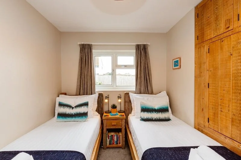 A small bedroom with two single beds separated by a wooden nightstand, a window with beige curtains behind the beds, and a large wooden wardrobe on the right wall.