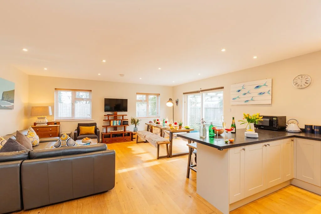 Bright open-concept living room and kitchen with wooden flooring, gray leather sofa, armchair, wall-mounted TV, dining table with chairs, and kitchen counter with appliances and decorations.