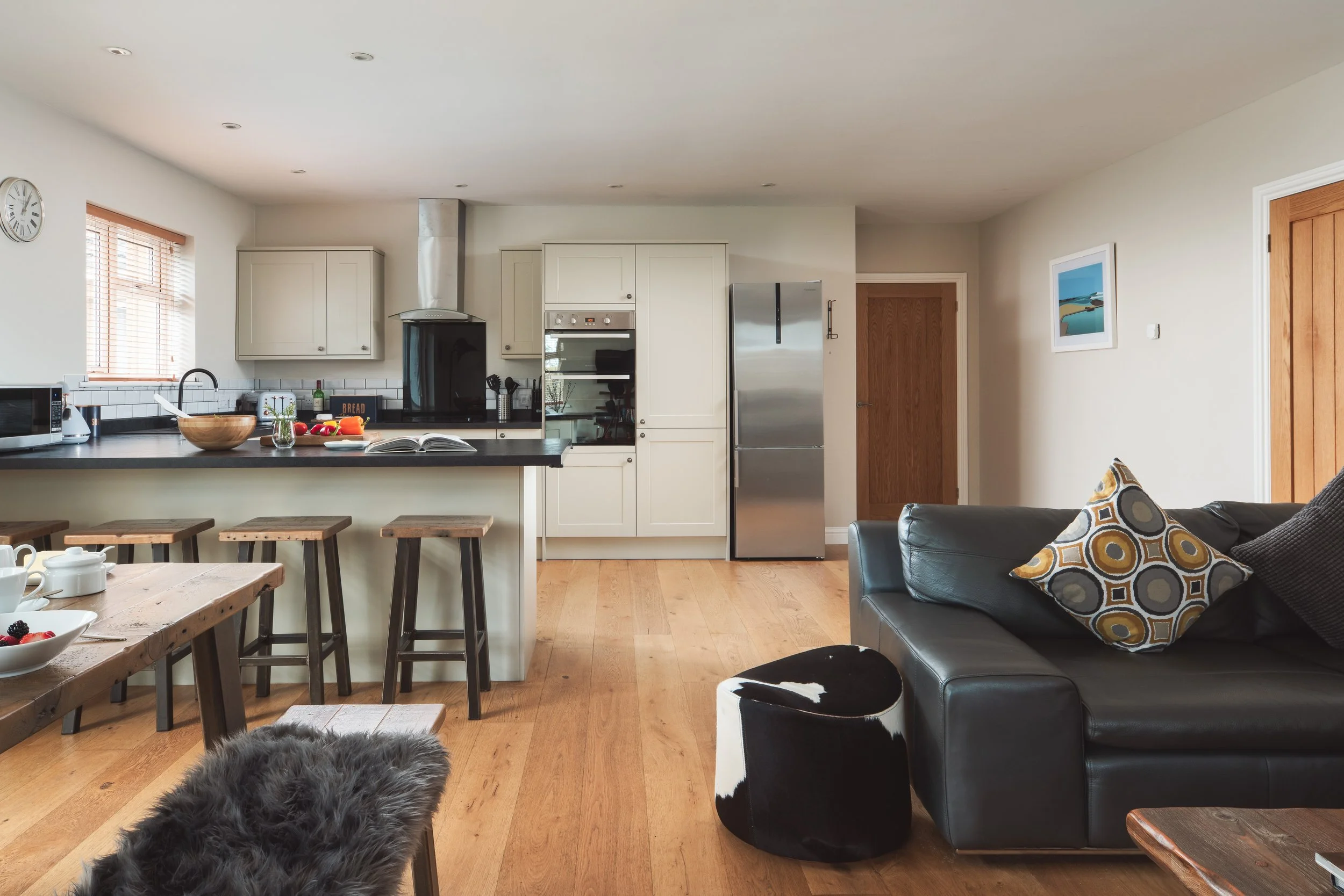 A modern kitchen and living room with white cabinets, stainless steel appliances, a black countertop island with three wooden bar stools, a wooden dining table with white cups and bowls, a black leather sofa with patterned cushions, and a wall art pa