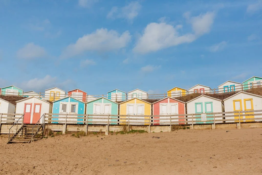 Colorful beach huts on a sandy shore under a blue sky with clouds, with stairs leading up to the huts.