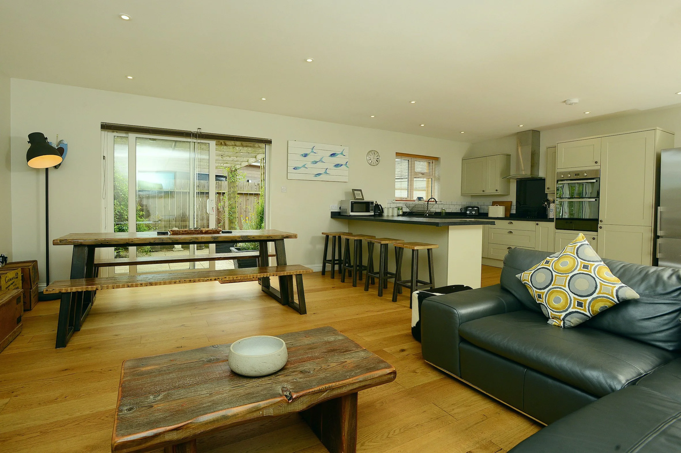 Open concept living room and kitchen with sliding glass door leading outside, wooden dining table, bar stools, black leather sofa with patterned pillows, artwork of fish, wall clock, microwave, oven, and stainless steel refrigerator.