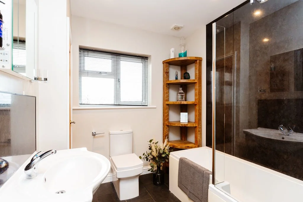 Modern bathroom with a white sink, toilet, window with blinds, wooden shelving unit, shower with dark glass, and decorative items.