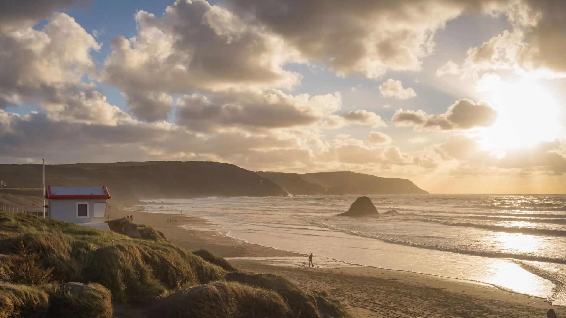A seaside landscape during sunset with a small white hut with a red roof in the foreground, a person walking along the beach, rocky formations in the water, a cliff in the background, and a sky filled with clouds.