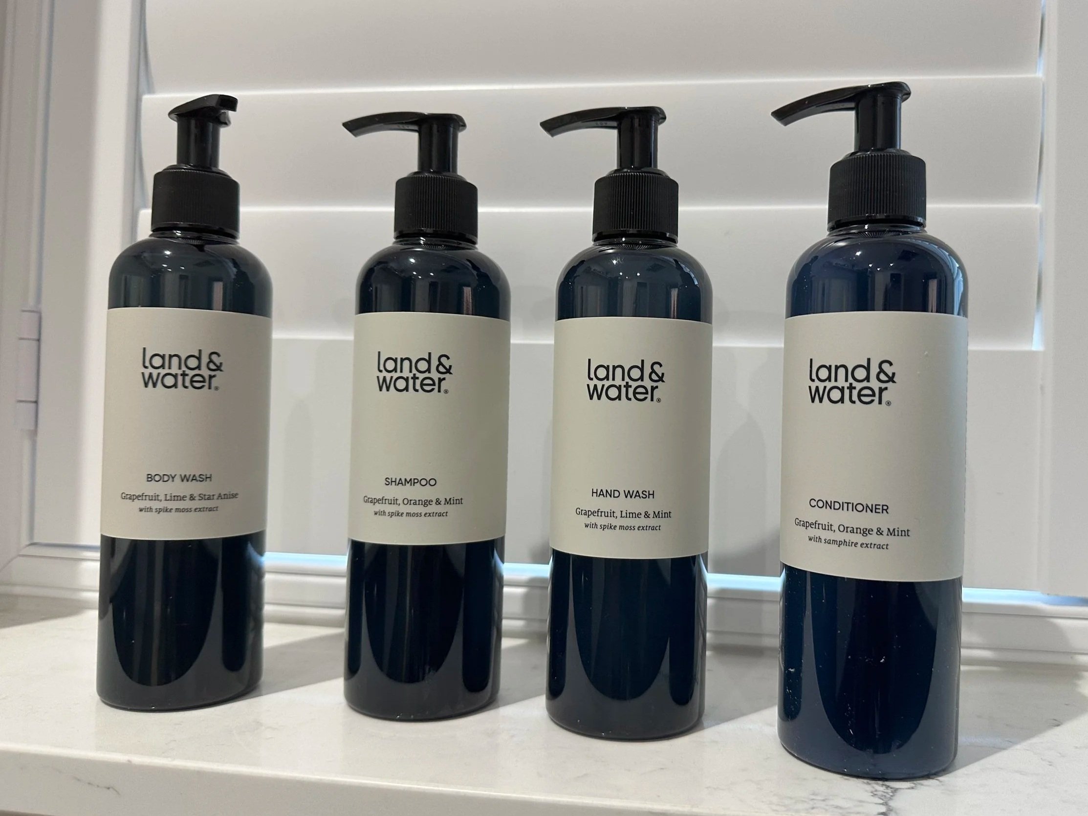 Four dark-colored bottles with pump dispensers labeled 'land & water', containing body wash, shampoo, hand wash, and conditioner, arranged on a white shelf.