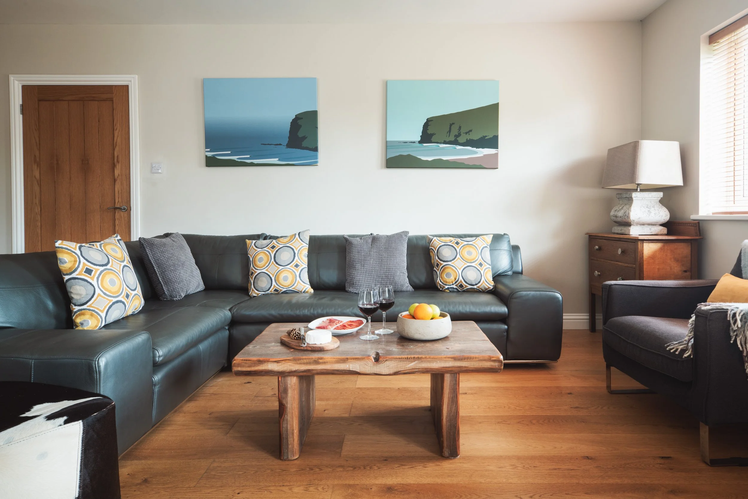 Living room with a dark leather sectional sofa, decorative pillows, wooden coffee table with snacks and drinks, wall art of ocean cliffs, and a window with blinds.