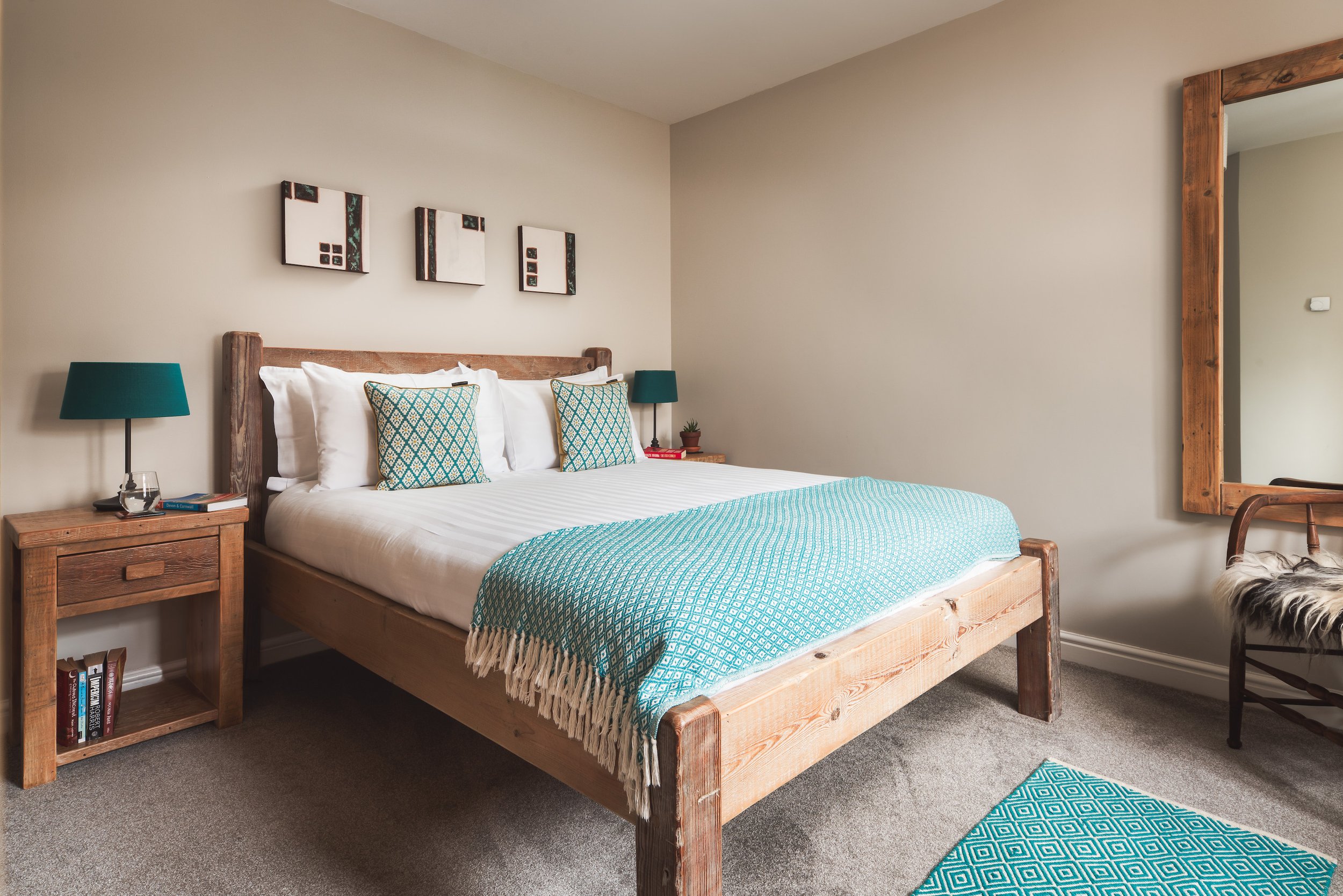 A bedroom with a wooden bed frame, white bedding, and patterned teal pillows. There is a wooden nightstand with a teal lamp, a small plant, and books. Three abstract art pieces are hanging above the bed. A mirror with a wooden frame is on the right w