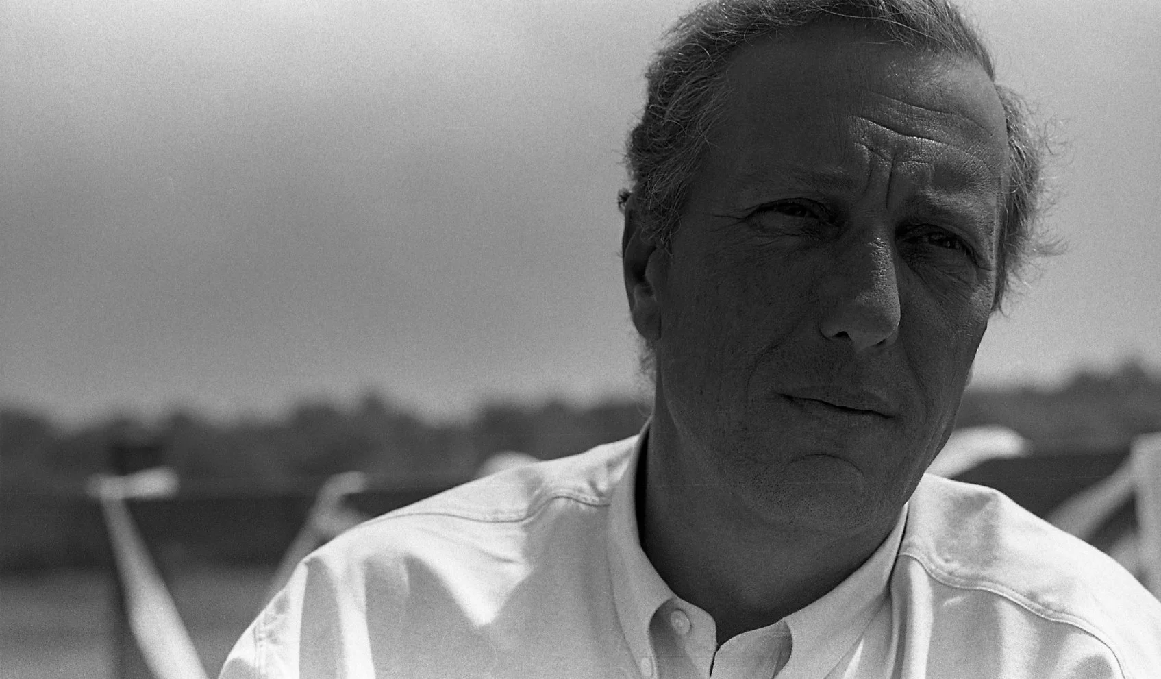 Close-up of a middle-aged man with grey hair, wearing a collared shirt, outdoors with boats and a body of water in the background, in black and white.