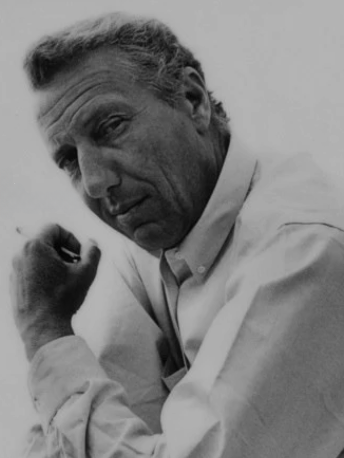 Black and white photo of a man posing with his fist near his face, wearing a button-up shirt, with a confident expression.