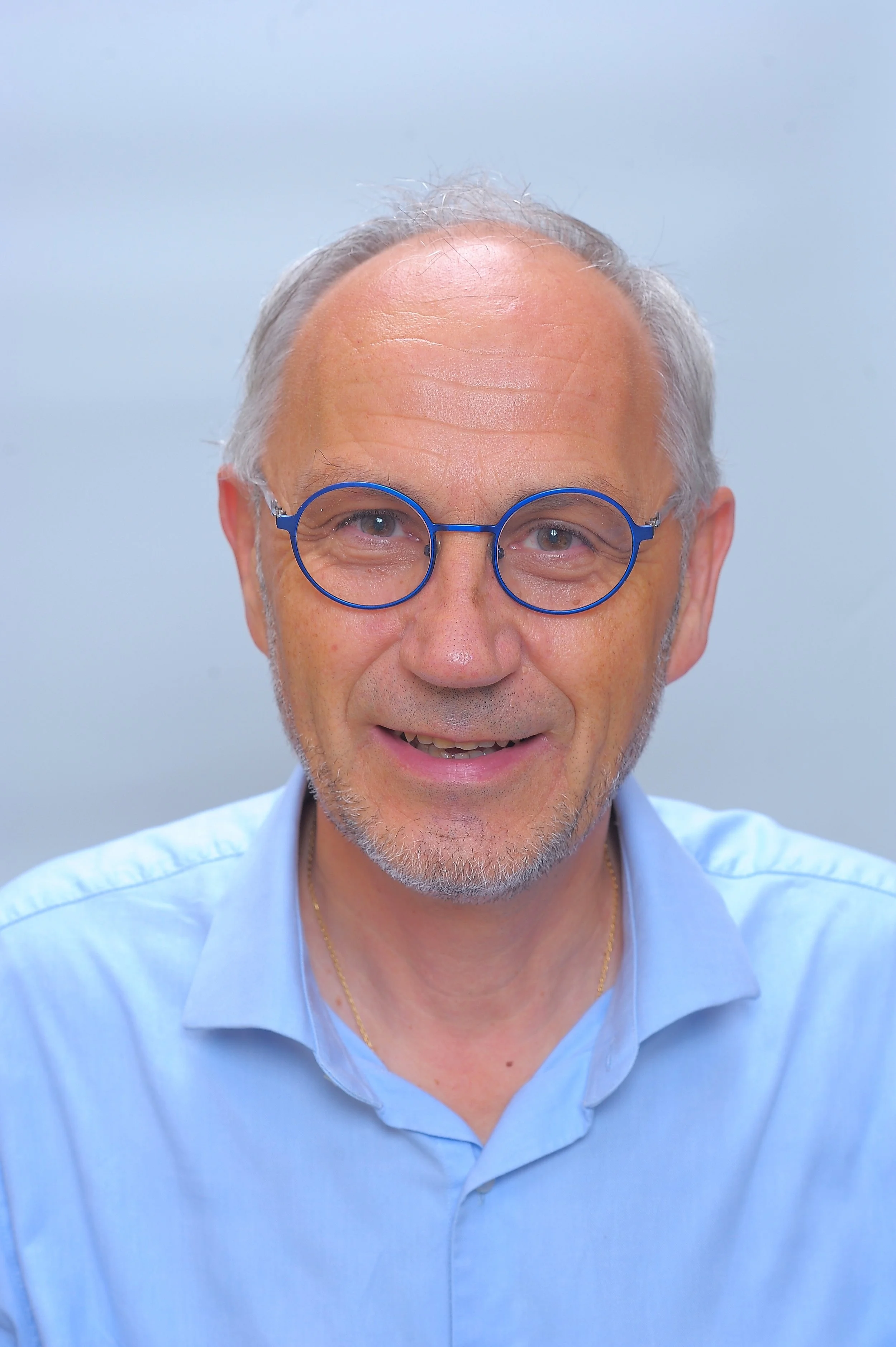 Un homme d'âge moyen avec des cheveux gris, portant des lunettes rondes bleues et une chemise bleue, souriant doucement, contre un fond gris clair.