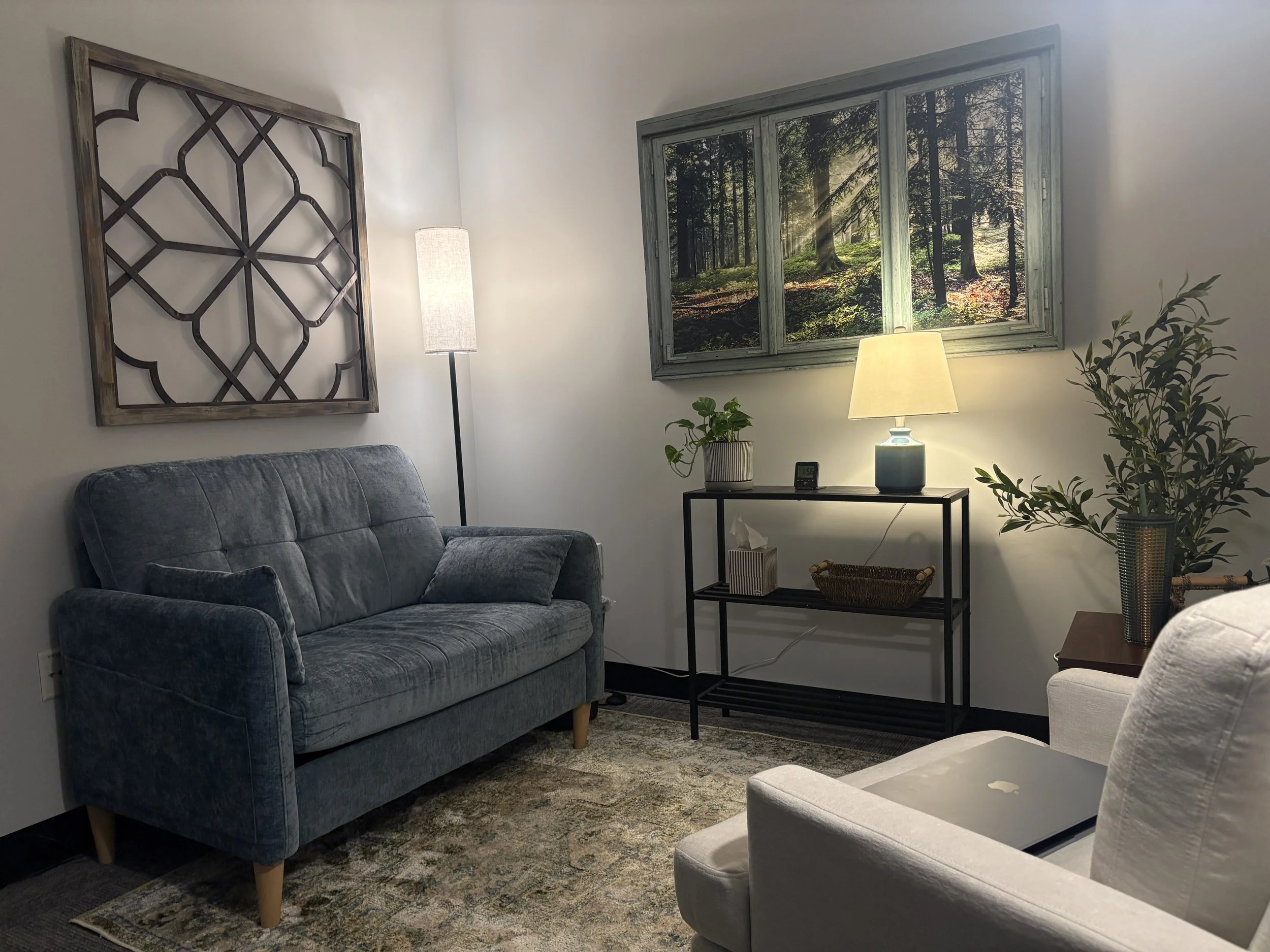 A cozy living room with a blue velvet sofa, a standing lamp, and a window with a forest view, decorated with houseplants and artwork.
