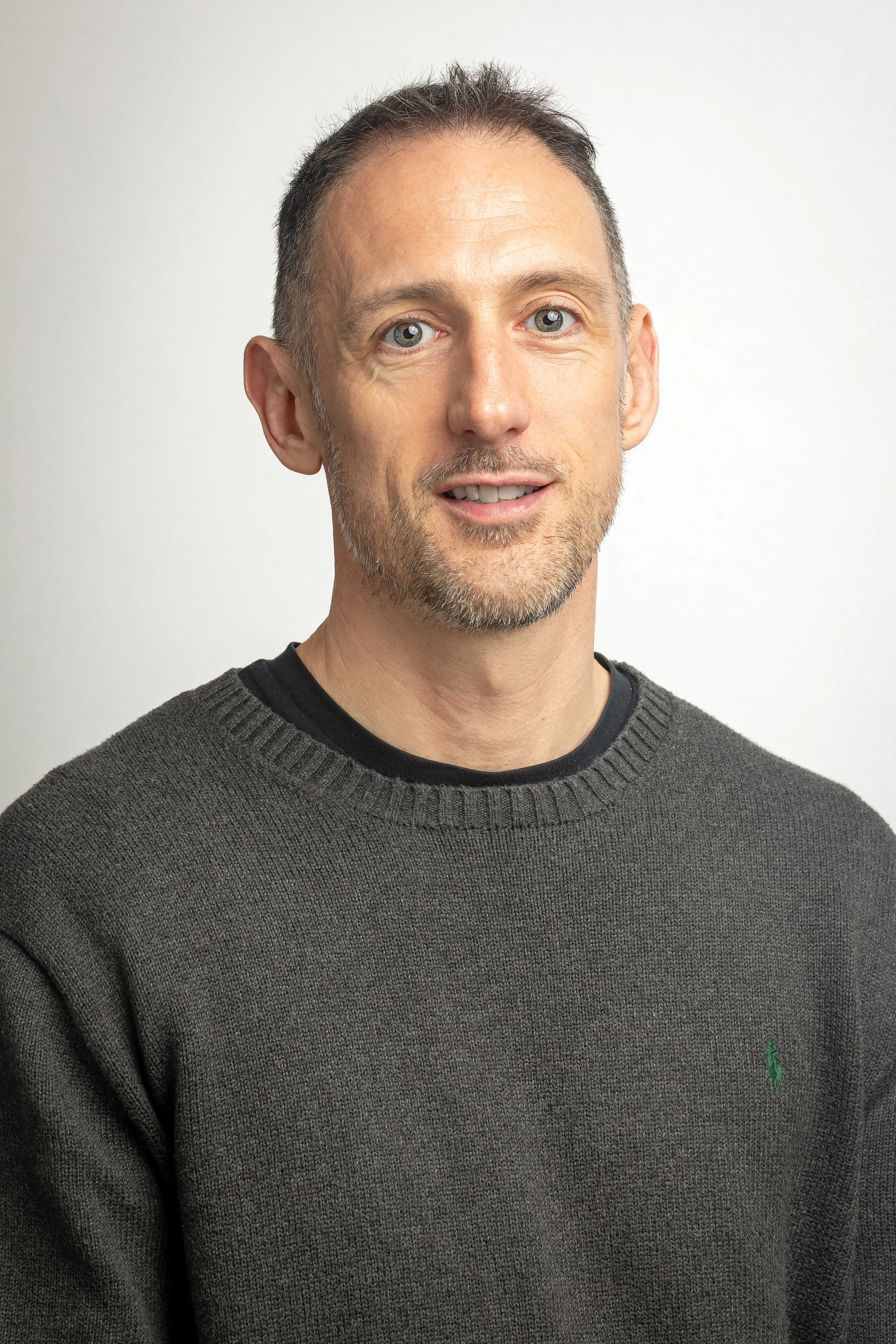 A middle-aged man with short dark hair, blue eyes, and a light beard, wearing a dark gray sweater with a small green logo, standing against a plain light-colored background.