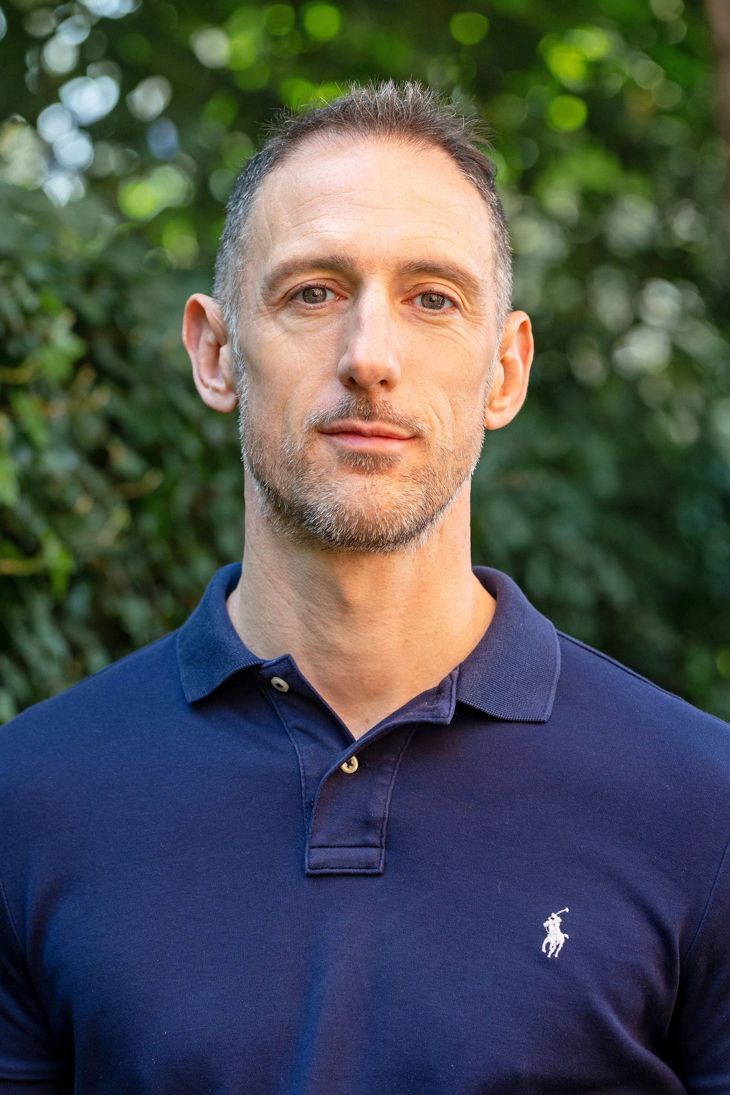 A middle-aged man with short gray hair and a beard, wearing a navy blue polo shirt with a small embroidered logo of a horse and rider on the left side, standing outdoors with a background of green trees and foliage.
