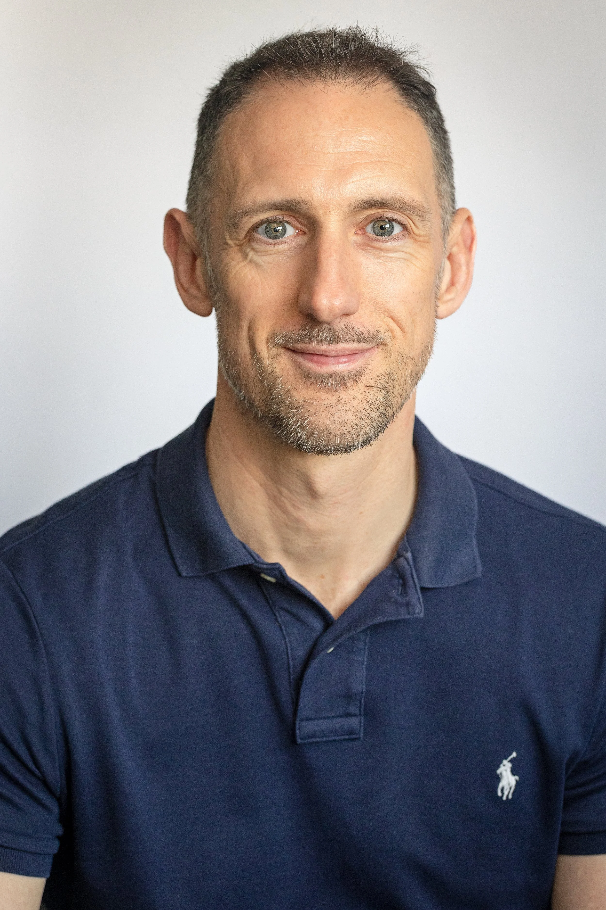A middle-aged man with short, gray hair and a beard, wearing a navy blue polo shirt with a small white logo of a polo player on the chest, smiling at the camera against a plain light gray background.