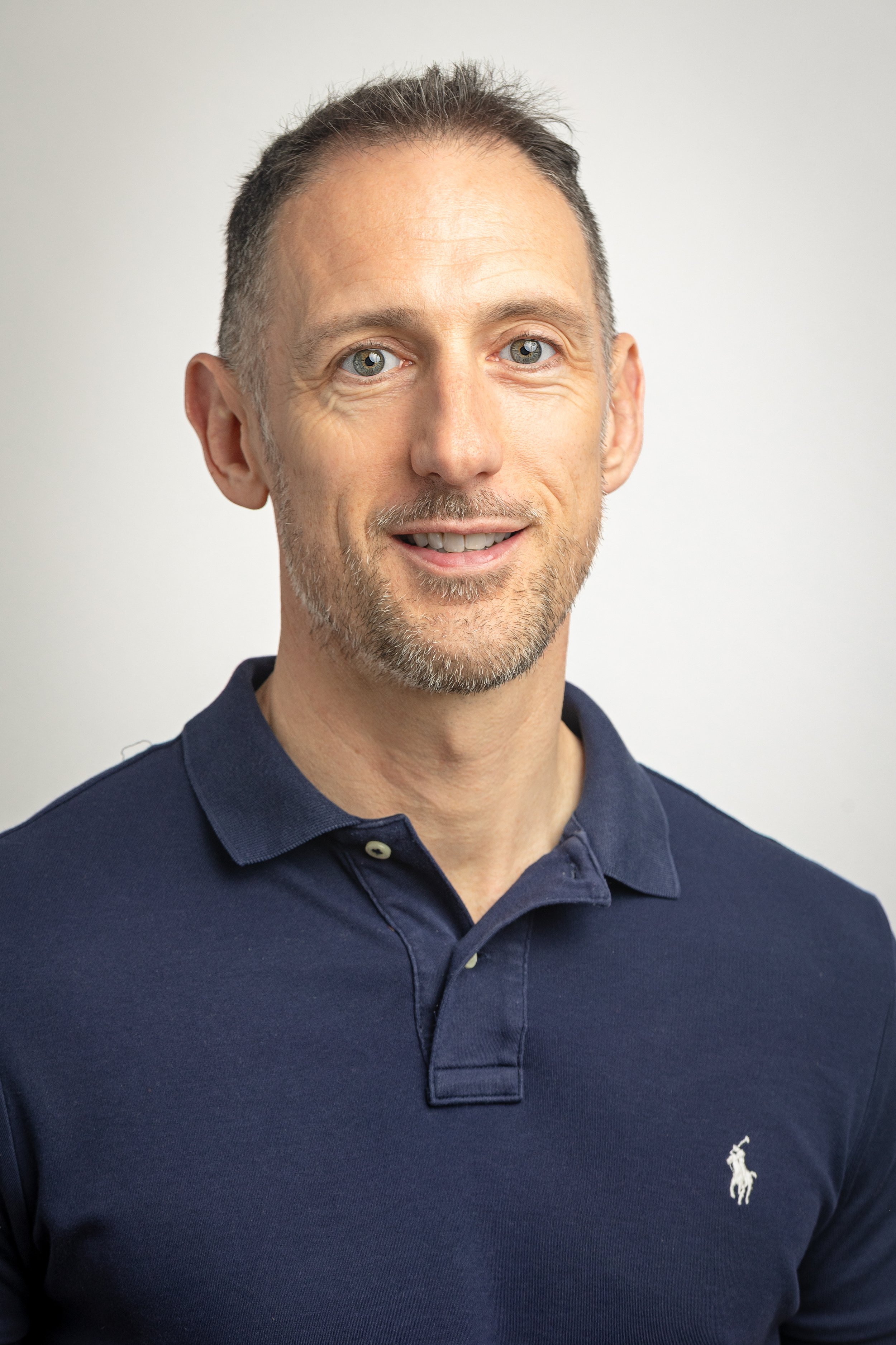 A middle-aged man with short dark hair, light skin, and a beard, wearing a navy blue Polo Ralph Lauren shirt, smiling against a plain white background.