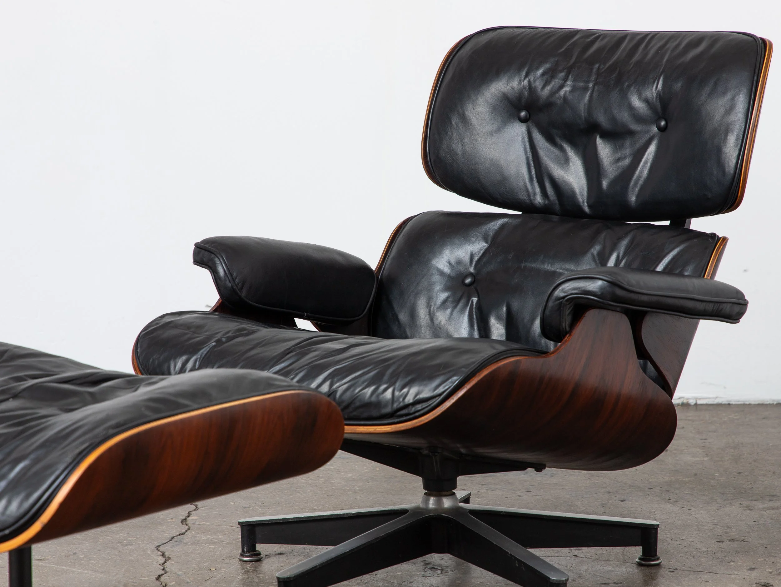 A black leather lounge chair with wooden accents and a matching ottoman, set against a plain white wall and visible concrete floor.