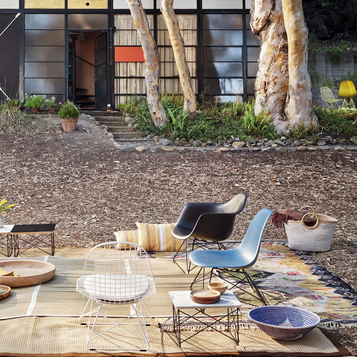 Outdoor patio area with modern chairs, a patterned rug, and a basket, set up on a dirt ground with trees and a building in the background.