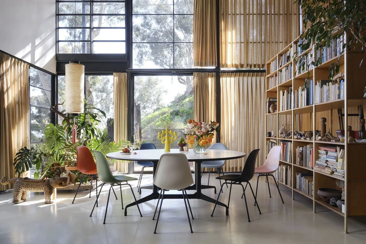 A bright modern room with large windows, a round dining table with colorful chairs, a bookshelf, plants, and decorative items.