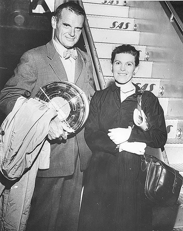 A man and a woman standing together on airport stairs, smiling at the camera. The man is holding a briefcase and a circular object, and the woman has a purse over her shoulder.